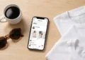 Smartphone displaying the Poshmark affiliate program dashboard on a wooden desk next to coffee and folded clothing, representing fashion reseller income.