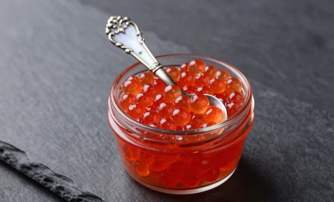 Glass jar of fresh red caviar with a mother-of-pearl spoon on a dark slate surface.