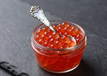 Glass jar of fresh red caviar with a mother-of-pearl spoon on a dark slate surface.