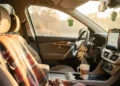 A warm and cozy car interior with plaid blanket draped over the seat, string lights on headrest, coffee in cup holder, and smartphone mounted on dashboard showing GPS navigation.